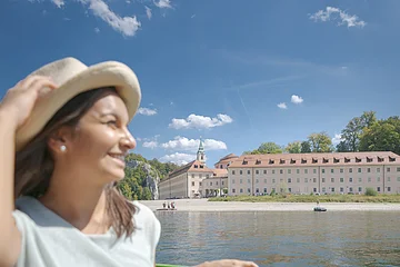 Eine junge Frau mit Strohhut und sommerlicher Kleidung sitzt in einem Ausflugsschiff mit gr&uuml;ner Sitzbank. Neben ihr liegt die Sonnenbrille auf der Bank. Im Hintergrund ist die Donau und das Kloster Weltenburg zu sehen. Am hellblauen Himmel sind einige wei&szlig;e Sch&auml;fchenwolken zu sehen.